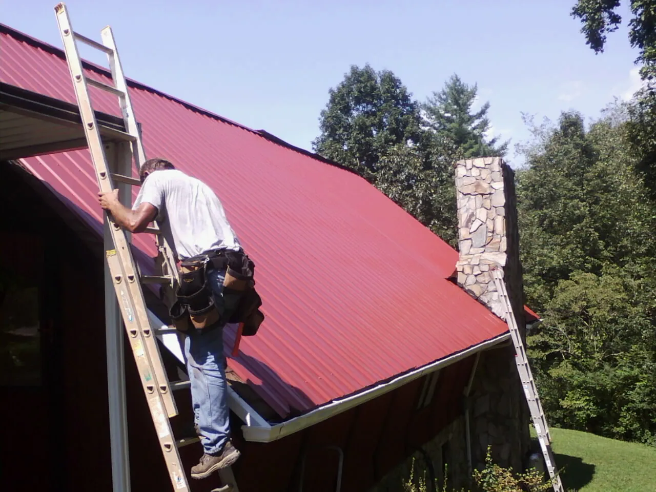 Roofer on a ladder beside a red metal roof and stone chimney — Soffit & Fascia Repair by Roofing Brothers in Lawrenceville